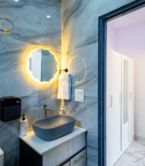 Bathroom with a white toilet, health faucet, vanity with grey basin and lit round mirror on grey tiled walls