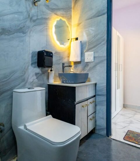 Bathroom with a white toilet, health faucet, vanity with grey basin and lit round mirror on grey tiled walls