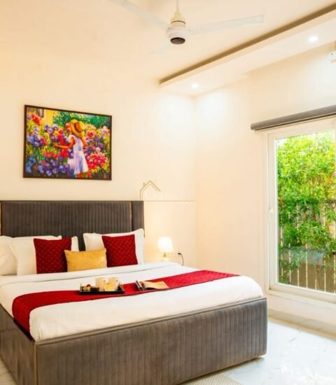 Bedroom with a grey bed, red cushions, a floral painting and a large sliding window facing green hedges