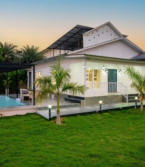 Farmhouse porch with a teal door and white railing lit at dusk beside the pool and garden path lights